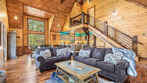 A spacious living room with a high ceiling and wooden walls, featuring two dark leather sofas with fur throw pillows, a wooden coffee table, and a staircase leading to an upper level.