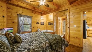Une chambre à coucher située dans une cabane en bois, avec un lit, un ventilateur de plafond, une télévision et une salle de bain visible à travers une porte ouverte.