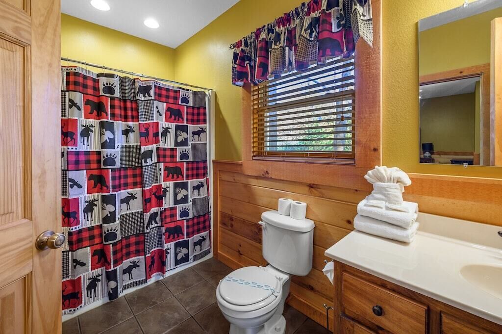 Un baño con cortina de ducha escocesa, un inodoro, un lavabo con espejo y una ventana con persianas.