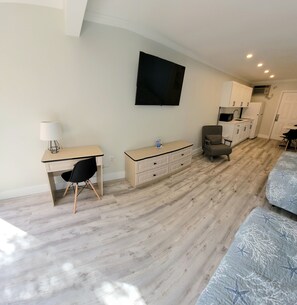 Una habitación moderna y bien iluminada con piso de madera, un escritorio con silla, un sofá, un televisor montado en la pared y una zona de kitchenette.