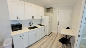 Una cocina compacta con armarios blancos, un microondas negro y un lavamanos. Hay una pequeña mesa con una silla, y una puerta blanca al fondo.