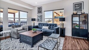 A spacious living room with a large black leather sofa, a wooden coffee table, and a herringbone-patterned rug. There are large windows offering views of the outdoors, and a black cabinet with glass doors on the right.