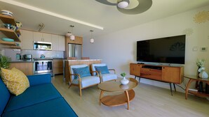 Una sala de estar moderna con sofá, sillas, una mesa de centro, televisor y una zona de cocina con electrodomésticos y estantes.