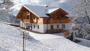 A wooden chalet with a balcony, a covered patio, and a snow-covered landscape.