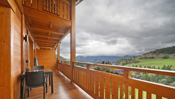 A wooden balcony with a table and chairs, overlooking a green landscape and mountains.