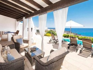 Ein überdachter Außenbereich mit Korbmöbeln, einem Tisch und Blick aufs Meer.