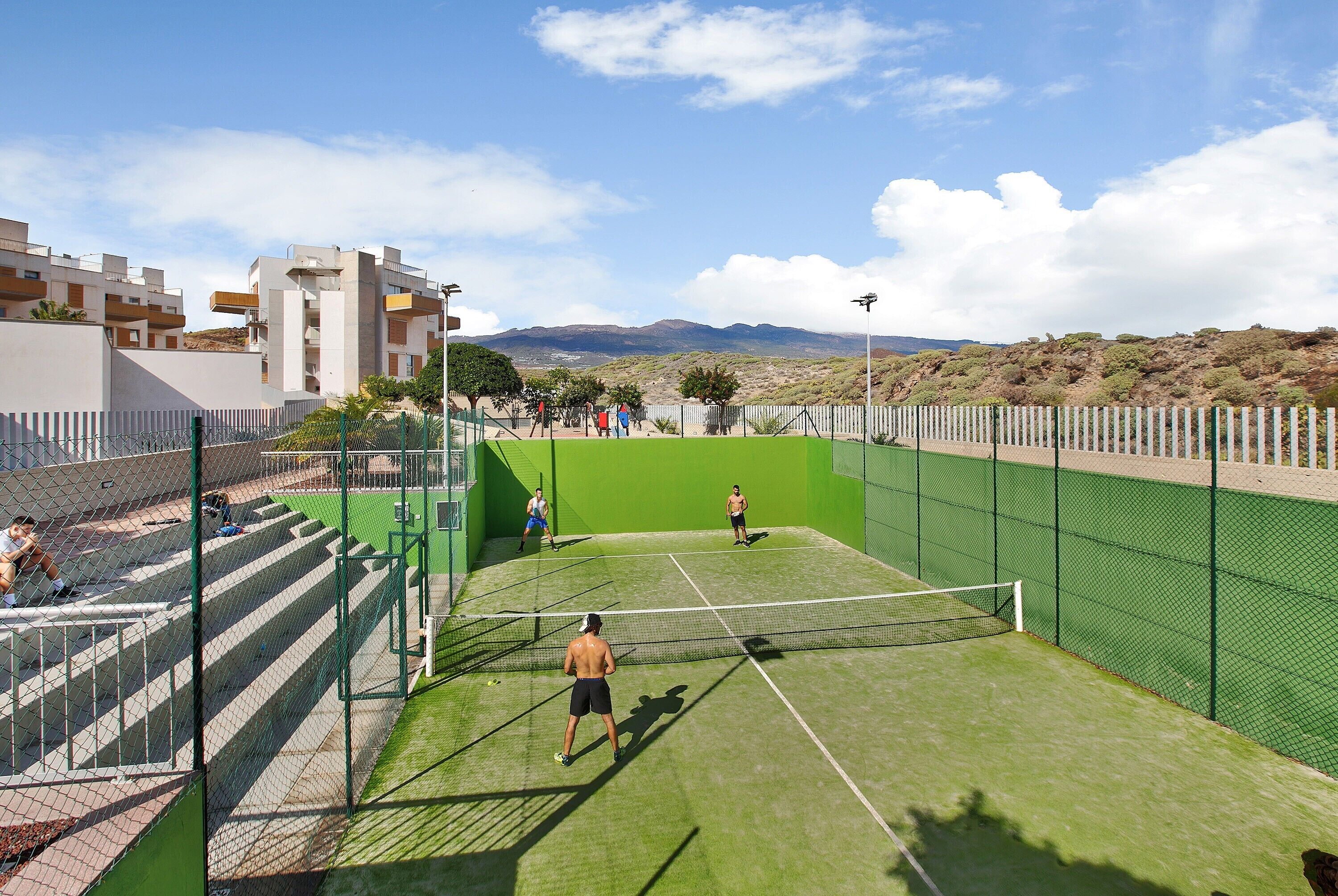 A tennis court with a net, surrounded by a green fence, and buildings in the background.