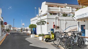 Een straat met een wit gebouw, genaamd 'Olympus Pool Bar', fietsen die er geparkeerd staan en een gele scooter.