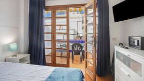 Een slaapkamer met een bed, een bureau, een stoel, een televisie en een schuifdeur naar een balkon.
