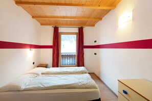 Un dormitorio con techo de madera, una cama con ropa blanca, una mesita de noche pequeña y una ventana con cortinas rojas.
