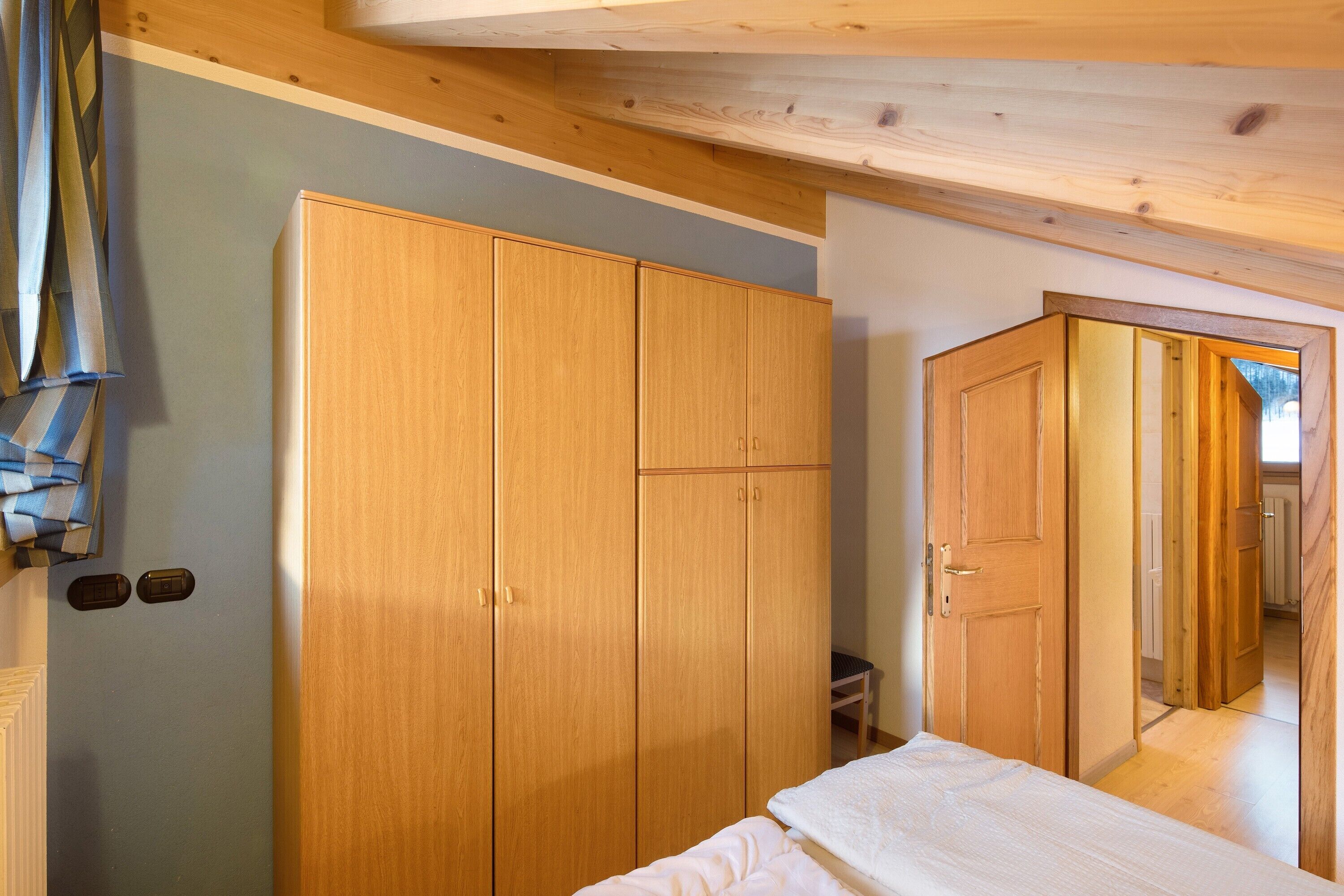 A bedroom with a wooden wardrobe, a bed, and a door leading to another room.
