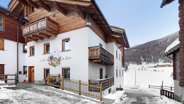 Un edificio de varios pisos con balcones de madera, suelo cubierto de nieve y montañas al fondo.