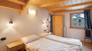 Un dormitorio con techo de madera, una cama con ropa de cama blanca, una mesita de noche, una ventana con vistas a un edificio de piedra y una puerta.