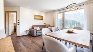 Una sala de estar moderna con una mesa de comedor de madera, un sofá y vistas a un bosque a través de amplios ventanales.