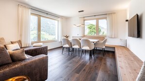 Una sala de estar moderna con sofá, mesa de comedor y sillas, ventanas amplias con cortinas, y un televisor de pantalla plana montado en la pared.