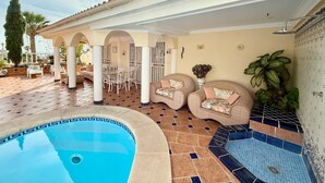 Un área de piscina con solárium de azulejos, mobiliario exterior y un porche cubierto.