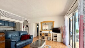 A living room with a blue sofa, a glass coffee table, a TV, and a kitchen area with cabinets and a refrigerator. - Pet Friendly Villa Azul close to the Beach with Pool, Garden, A/C and Wi-Fi (Callao Salvaje, Tenerife)