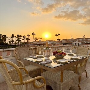 A dining table set for four with wicker chairs, a fruit bowl, and a bottle of champagne.