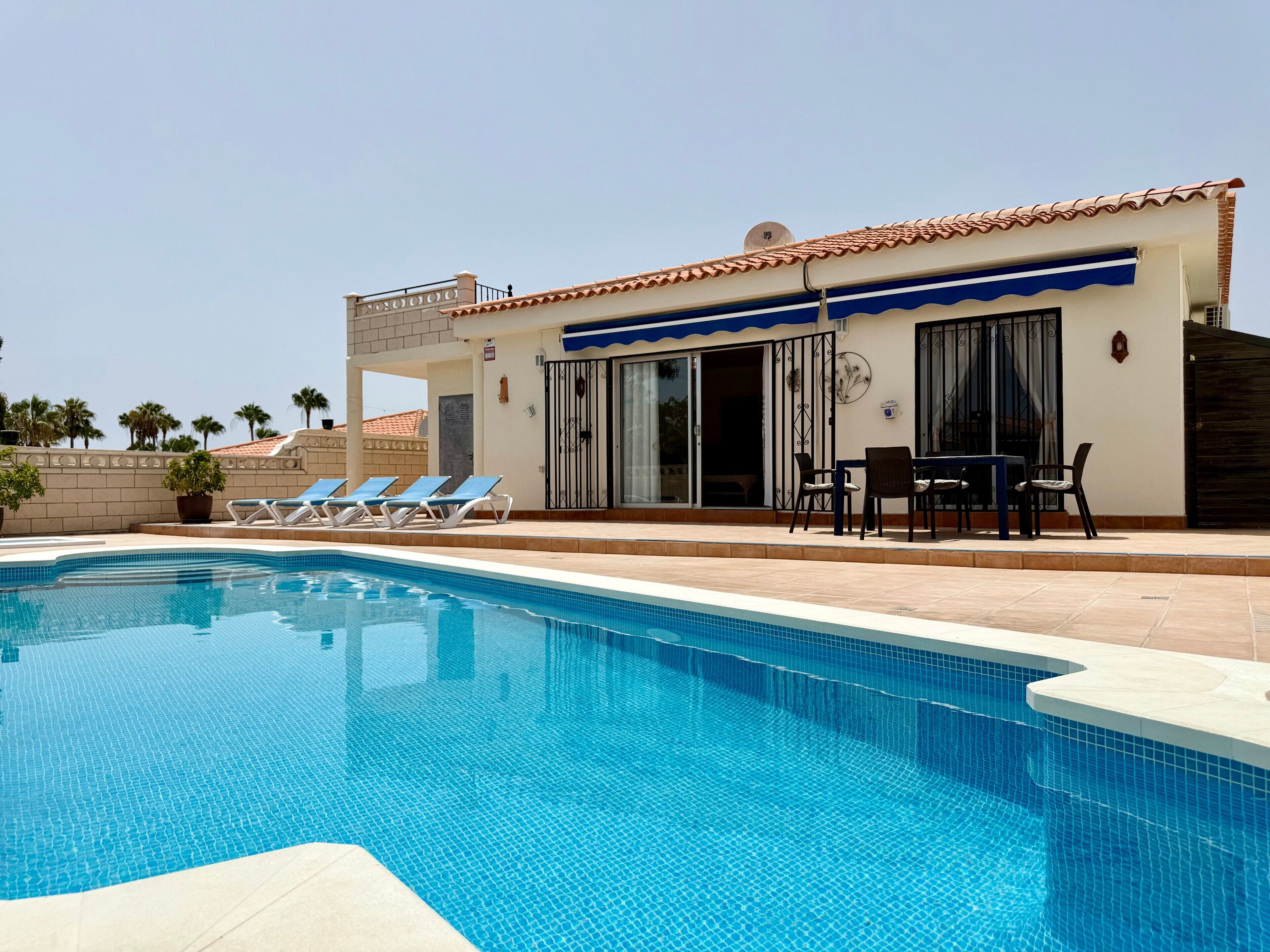 Una piscina con sillas de descanso, una mesa de patio y un edificio con techo de tejas y puertas correderas de vidrio.