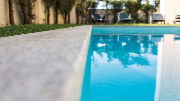 Una piscina con superficie de agua azul cristalina, rodeada por un deck de concreto y un muro bajo con árboles.