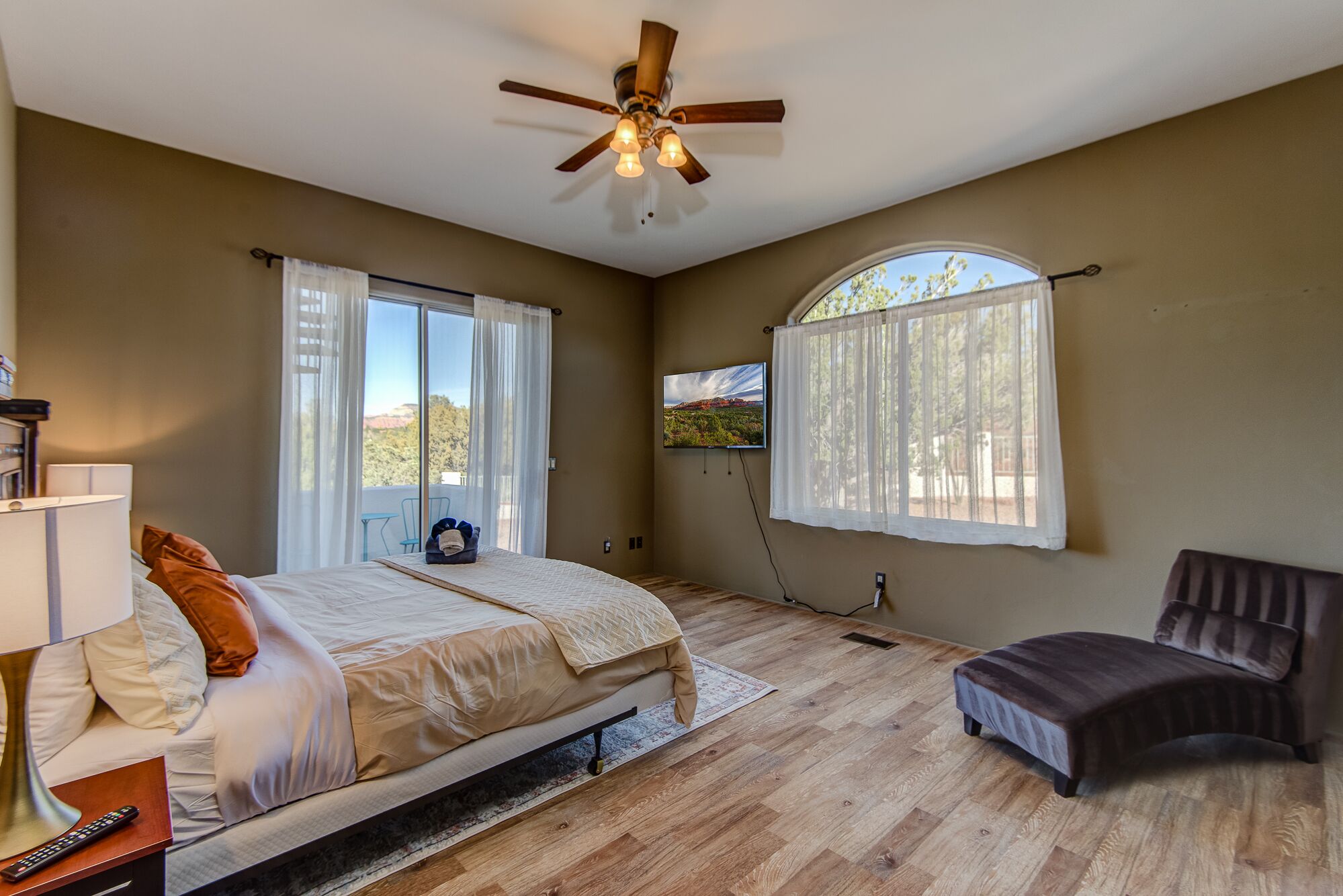 A bedroom with a bed, a desk, a chair, a ceiling fan, a TV, and a window with curtains.