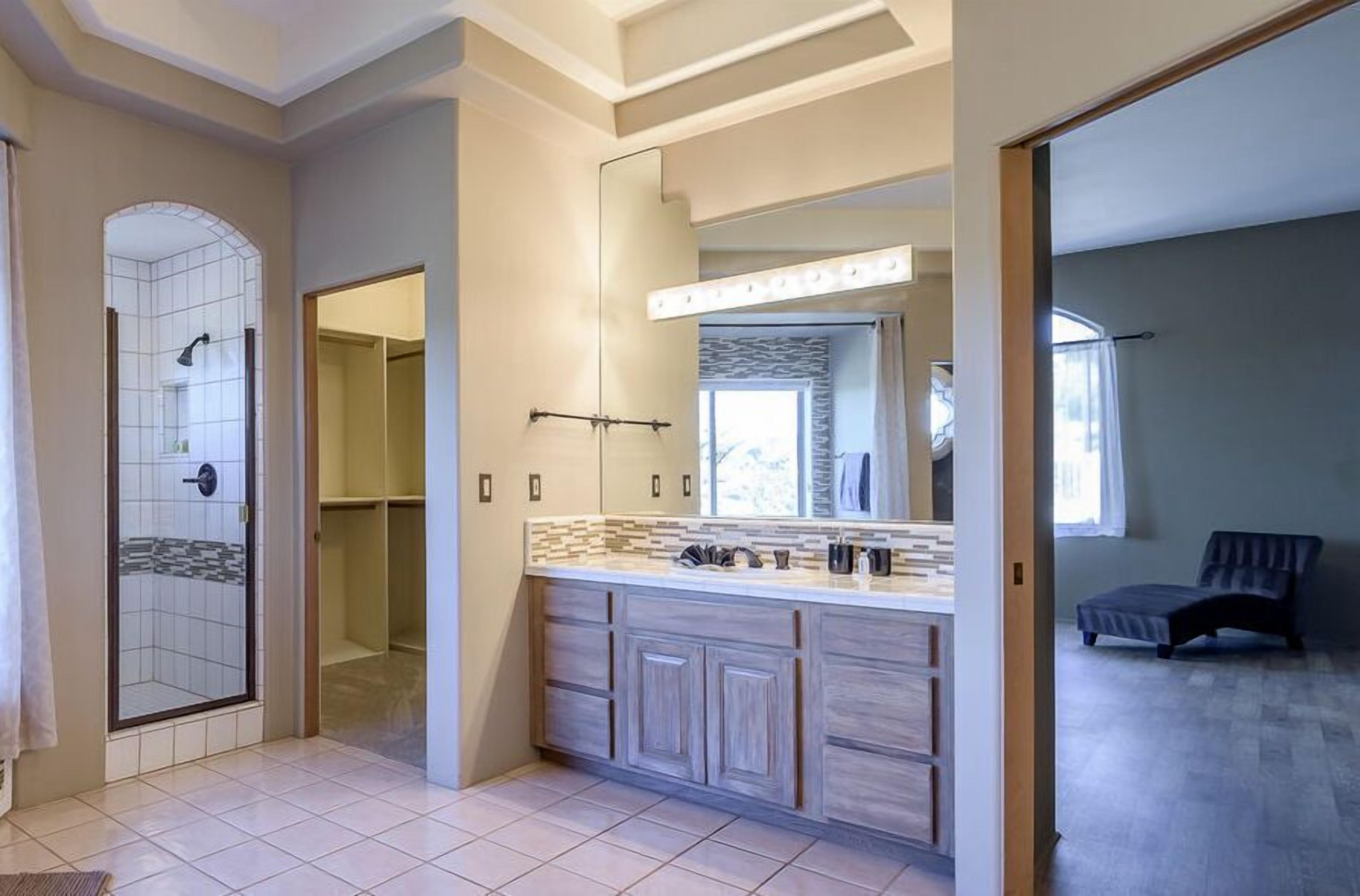A modern bathroom with a large vanity, a walk-in shower, and a separate bedroom area with a bed and a chair.