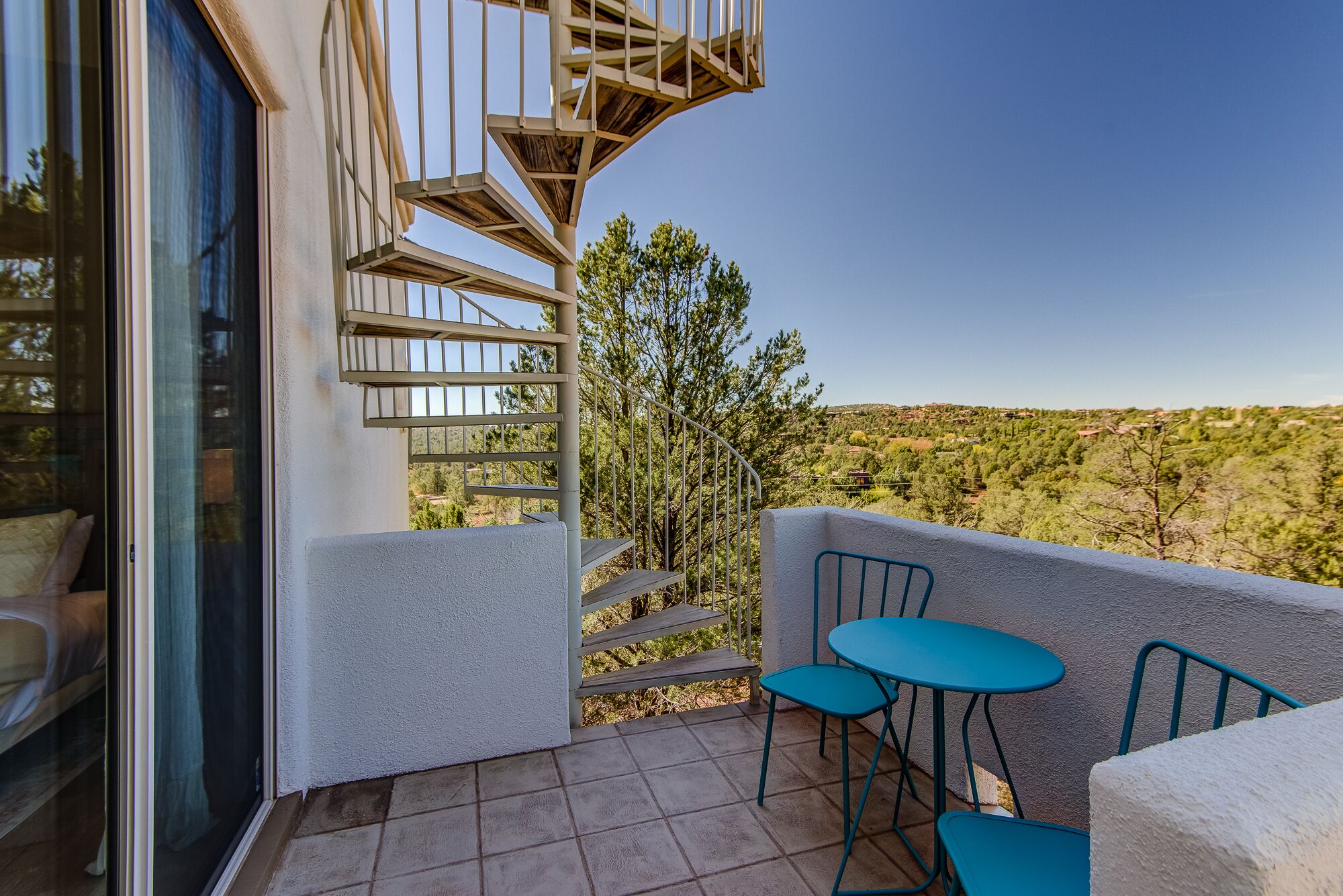 Ein Balkon mit Wendeltreppe, zwei blaue Stühle und Blick auf eine hügelige Landschaft.