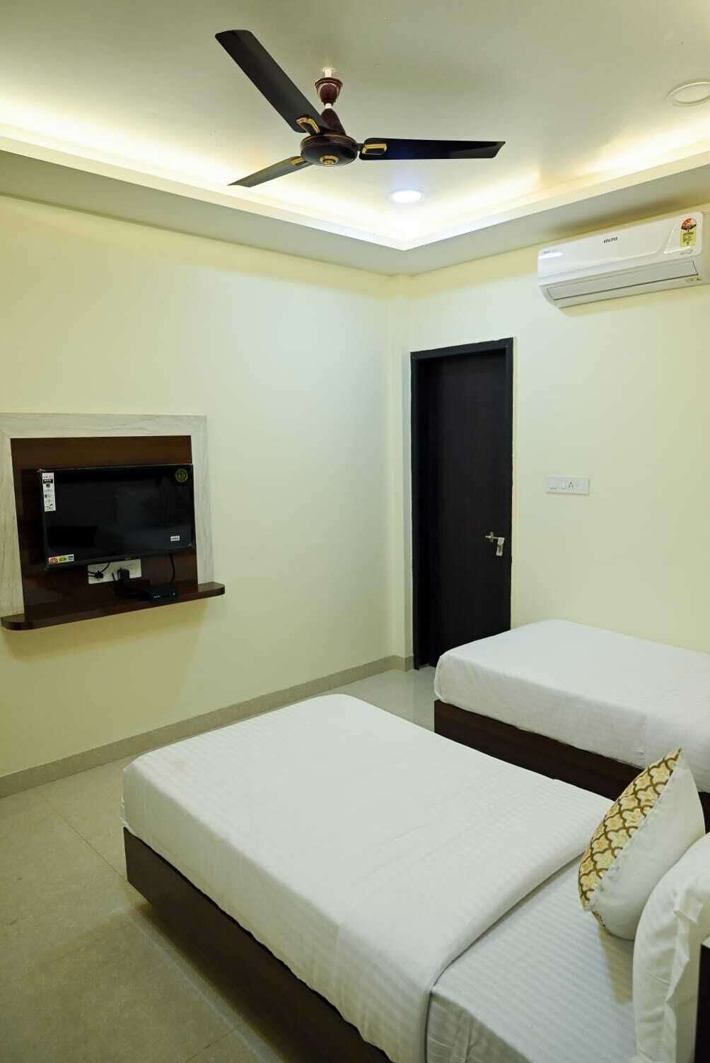 A hotel room with two single beds, a ceiling fan, a mounted TV, and an air conditioning unit.