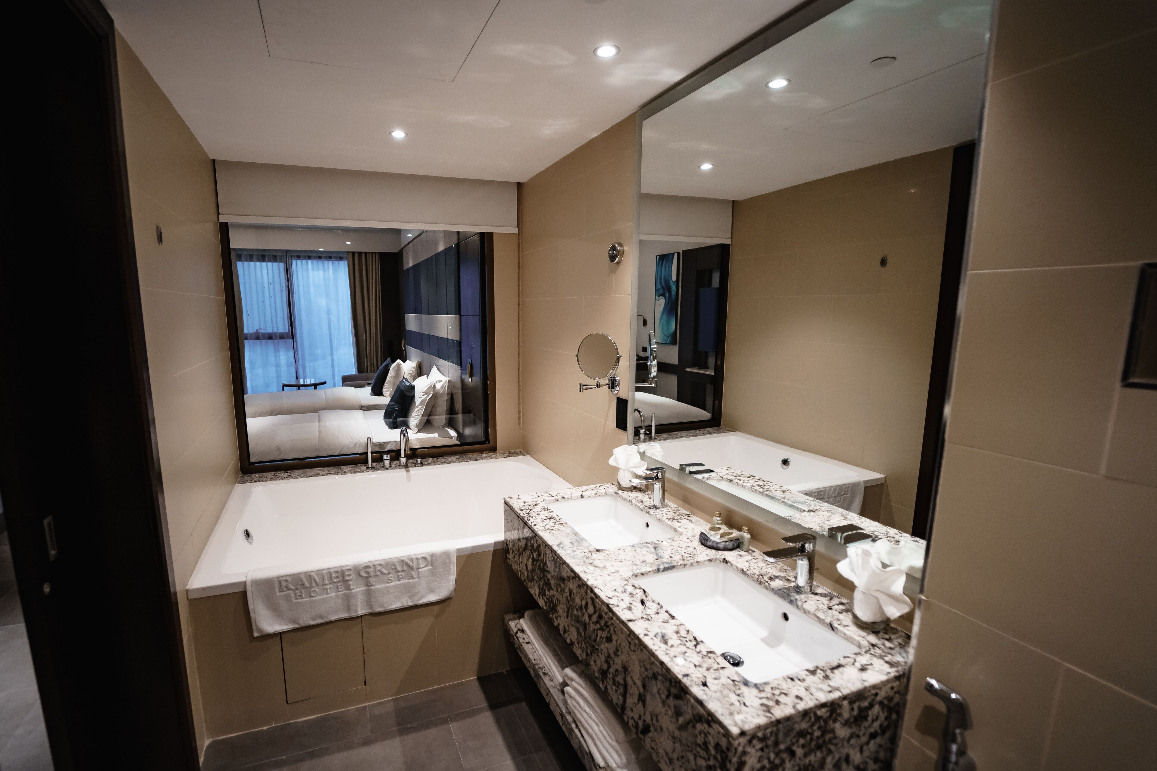 Un baño moderno con dos lavamanos, un espejo grande y vista a una habitación de hotel a través de un separador de vidrio.