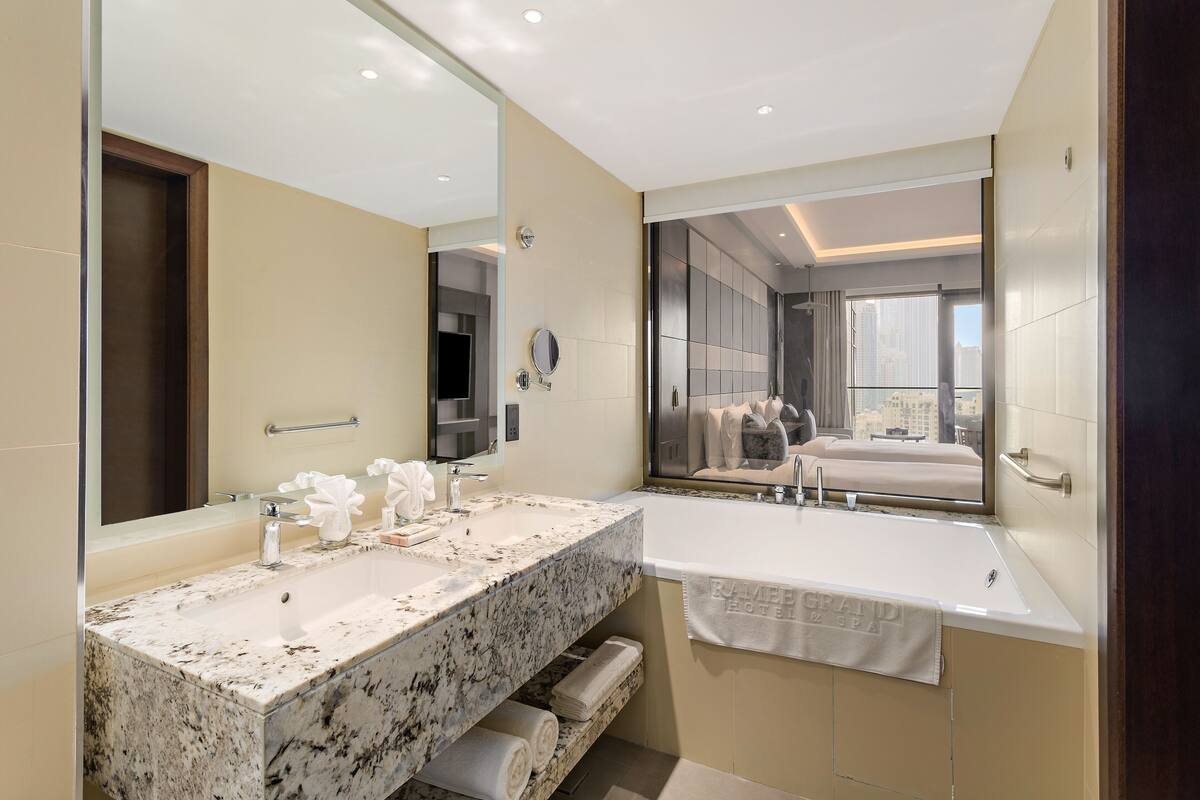 A modern bathroom with a large mirror, a granite countertop, a bathtub, and a view of the city through the window.
