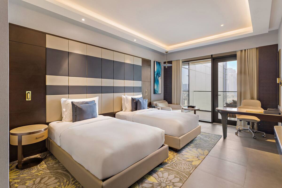 A modern hotel room with two beds, a desk, and a large window offering a city view.