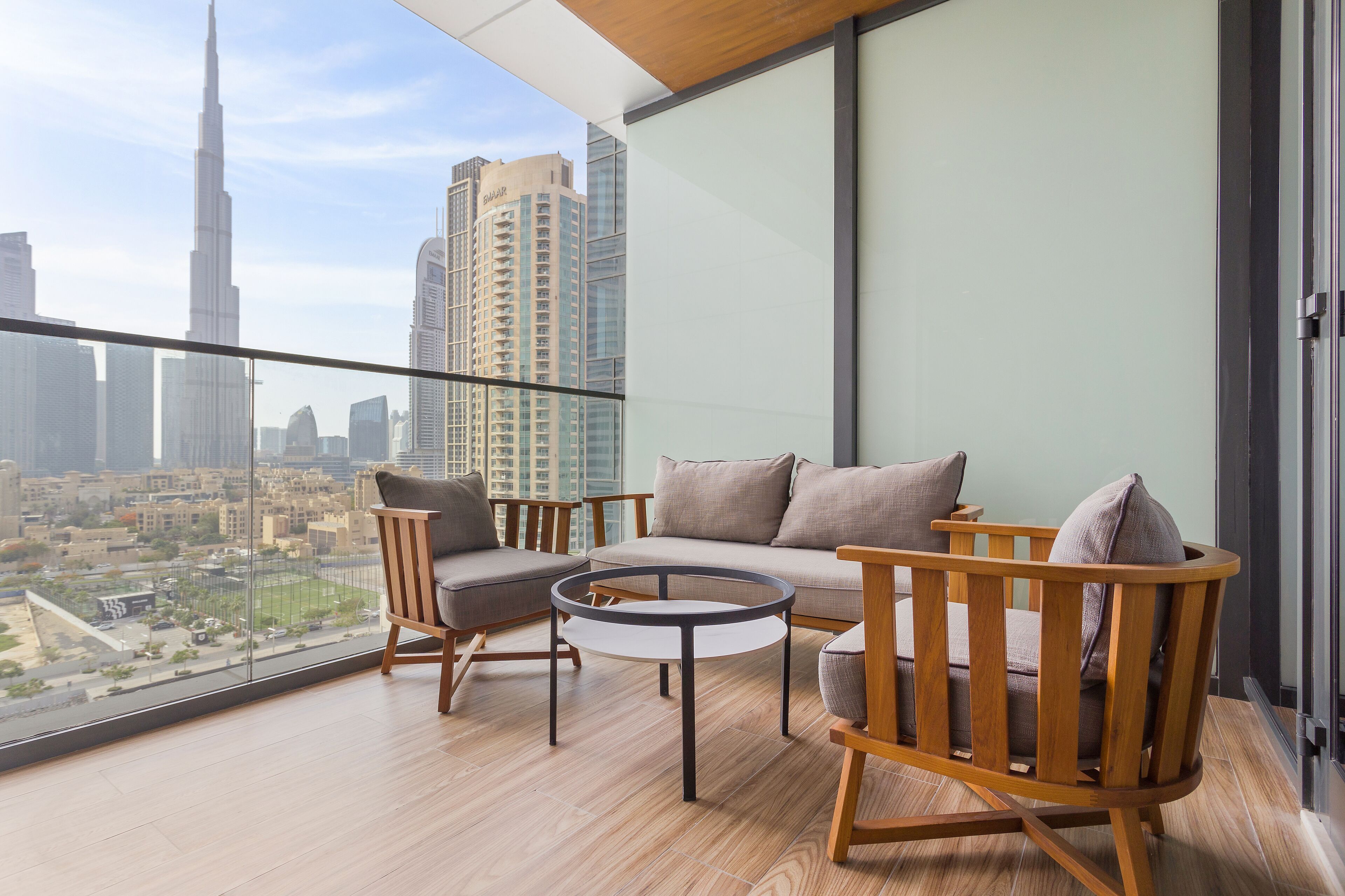 A balcony with a sofa, chairs, and a small table, overlooking a cityscape with tall buildings.