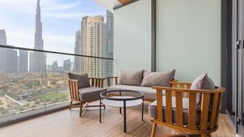 A balcony with a sofa, chairs, and a small table, overlooking a cityscape with tall buildings.