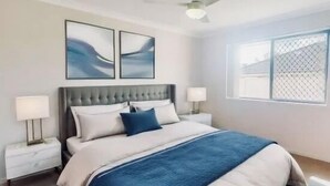 A bedroom with a bed, two bedside tables, a ceiling fan, and a window with a view. - Sandstone Point (SEQ) 4 bedroom house (Sandstone Point)