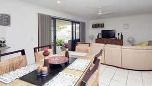 A dining area with a table set for four, a sofa, a TV, and a fan in the background. - Sandstone Point (SEQ) 4 bedroom house (Sandstone Point)