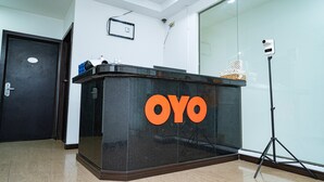 Un taulell de recepció amb el nom "OYO" en lletres negretes, una porta tancada, una paret de vidre i un trípode amb una càmera al costat dret.