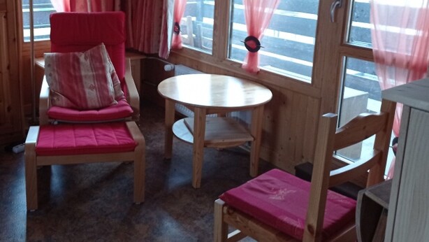 A cozy room with wooden walls and large windows, featuring a red chair, a small round table, and a wooden bench. - 2-room South/West with Wifi in Tignes, 5 sleeps, mountain view (Tignes)