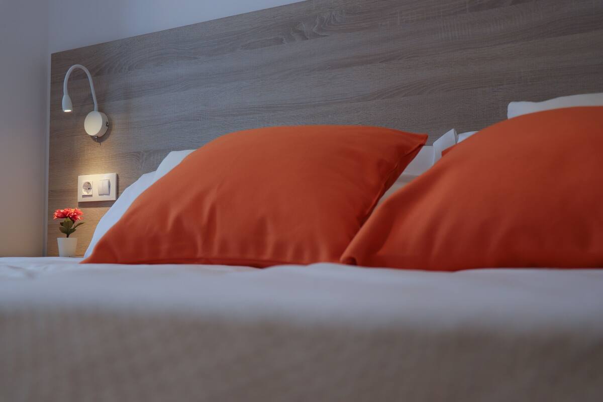 Ein Bett mit weißer Bettwäsche und zwei orangefarbenen Kissen, eine Wandleuchte und ein kleiner Blumenvasen.