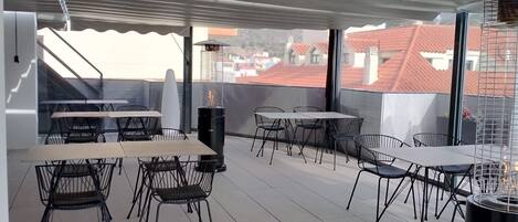 Una terraza en la azotea con mesas y sillas, una parrilla y vistas a edificios con tejados rojos.