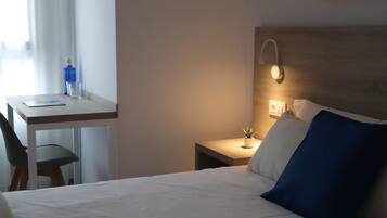 A hotel room with a bed, a desk, a chair, a nightstand, and a wall-mounted lamp.