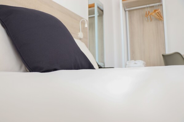 Habitación de hotel con cama, pared blanca, cabecero de madera, mesita de noche blanca, lámpara blanca y puerta de madera con perchero para toallas.