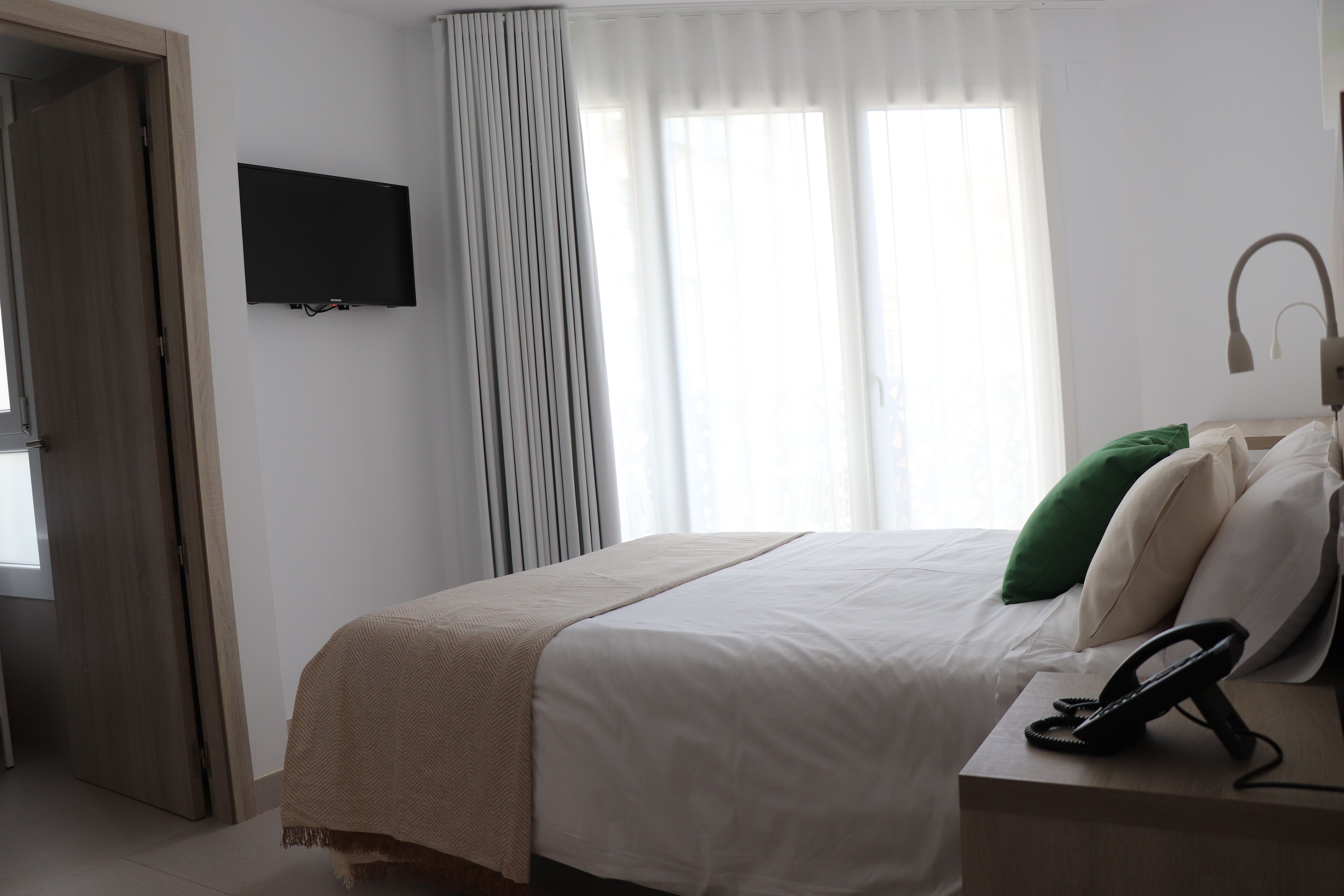Habitación de hotel con cama, teléfono en la mesita de noche, televisor en la pared y ventana con cortinas.