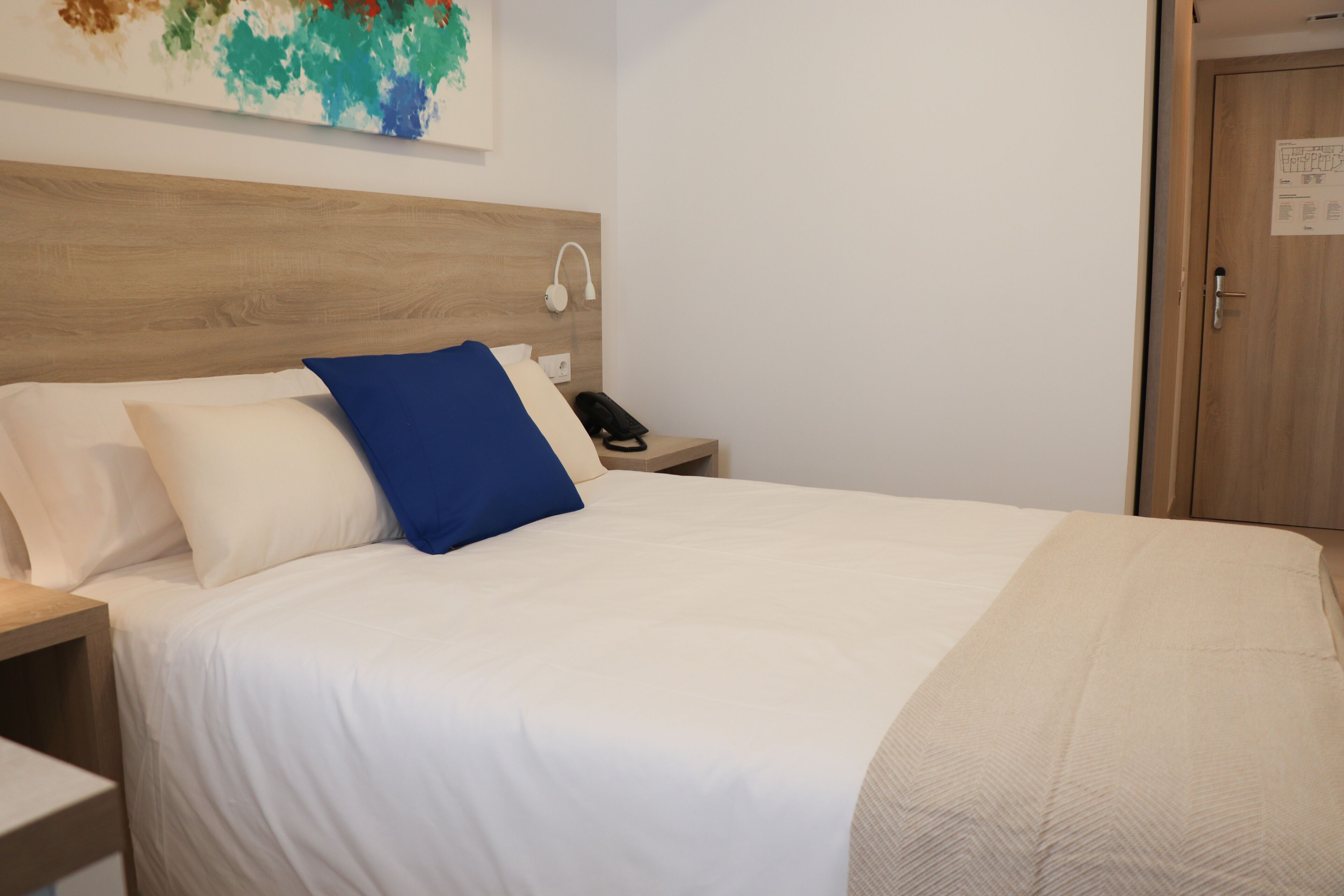 Uma cama bem arrumada com almofadas brancas e azuis, uma cabeceira de madeira, uma mesa de cabeceira com um telefone e uma obra de arte na parede acima da cama.