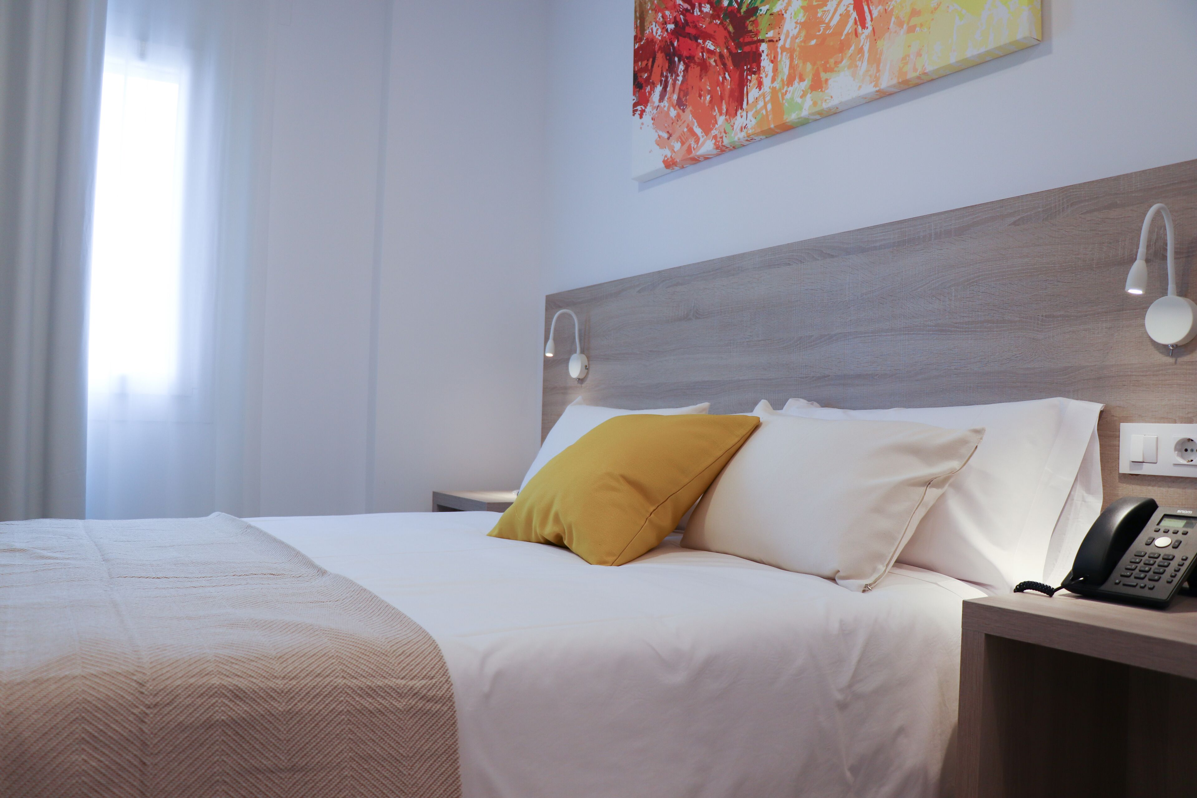 A hotel room with a bed, a bedside table with a telephone, and a painting on the wall.