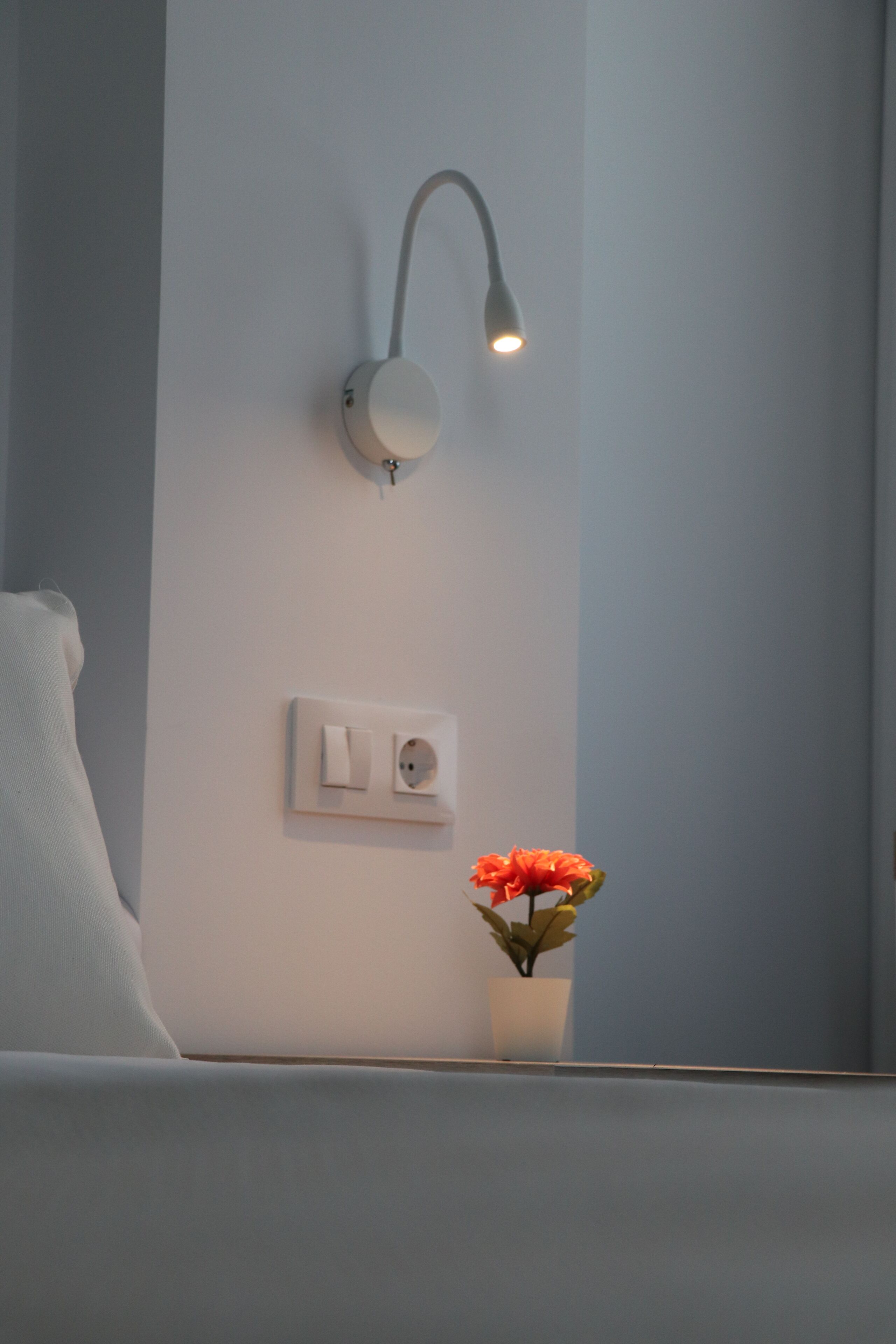Una habitación con pared blanca, mesita de noche blanca, lámpara blanca y una planta en maceta con flores anaranjadas.