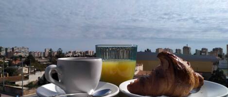Un desayuno con un croissant en un plato, una taza de café y un vaso de zumo de naranja sobre una mesa con vistas a la ciudad al fondo.