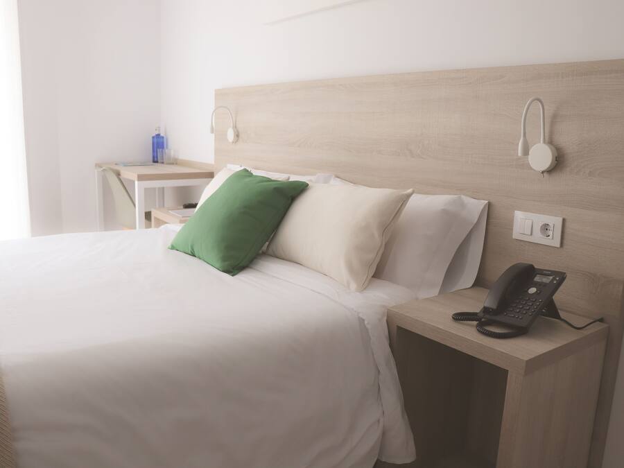 A neatly made bed with white bedding and a green pillow, a bedside table with a telephone, and a wall-mounted light fixture.