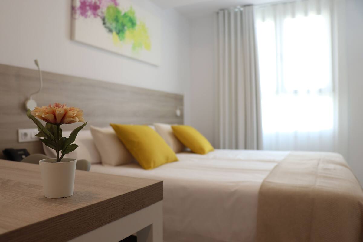 Ein Hotelzimmer mit Bett, Nachttisch mit Topfpflanze und Fenster mit Vorhängen.