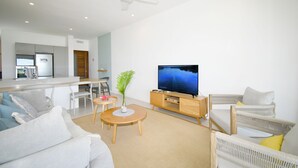 Uma sala de estar moderna com televisão de tela plana, ventilador de teto e área de jantar com cozinha ao fundo.