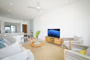 A modern living room with a flat-screen TV, a ceiling fan, and a dining area with a kitchen in the background. - Modern White and Blue on the beach with pool (Poste Lafayette)